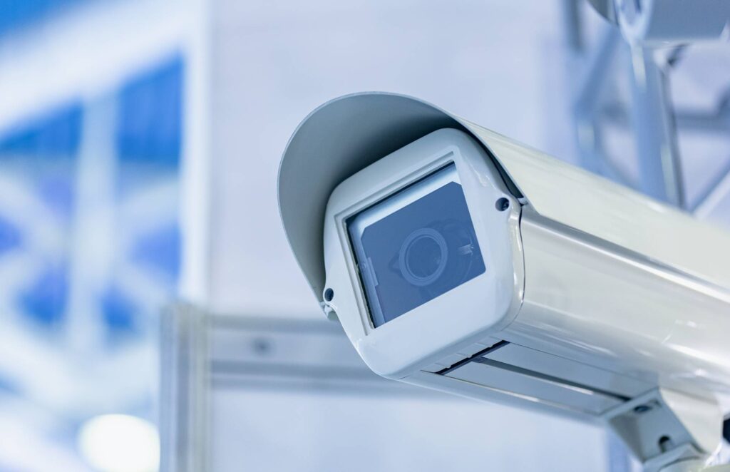 Close-up of a modern security camera with a white casing, mounted indoors, facing slightly downward. The background is blurred with blue and white tones.