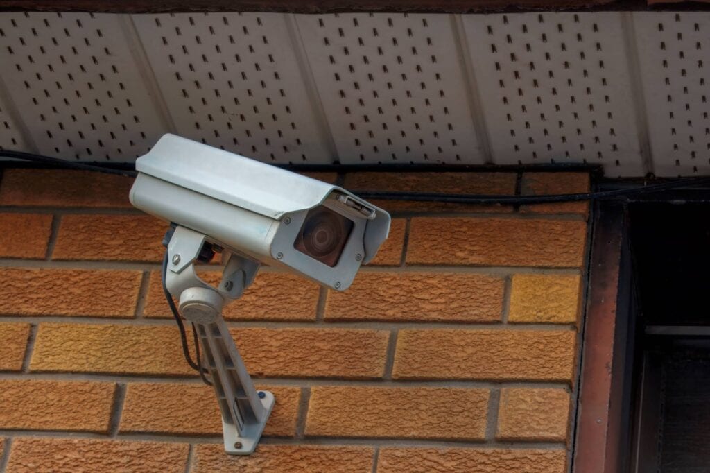 A security camera mounted on a brick wall, angled slightly downward, with a house roof overhang visible above.